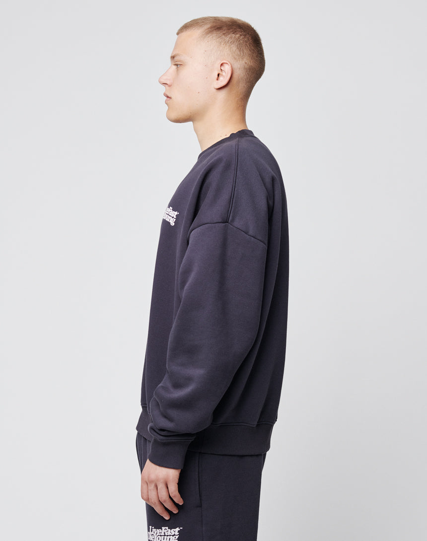 A person with short blond hair stands in profile against a plain background, wearing a dark LIVE FAST DIE YOUNG Basic Chest Sweater and matching pants featuring small white prints on the chest and leg.