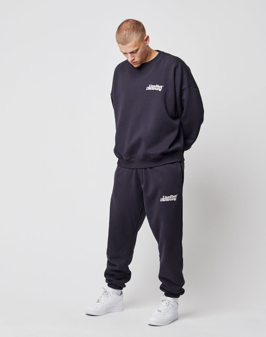 A person in the LIVE FAST DIE YOUNG Basic Chest Sweater, a loose-fitting black cotton piece with chest print, and matching sweatpants, pairs the look with white sneakers against a plain light gray background, gazing downward.