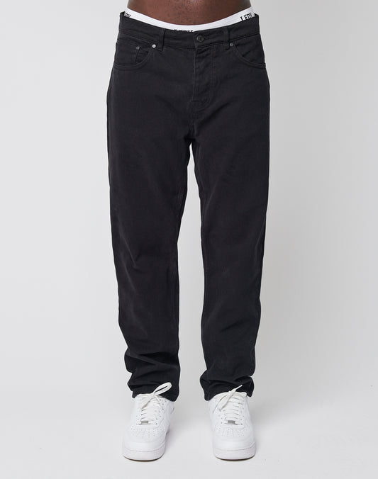 A person models LIVE FAST DIE YOUNG Regular Denim black jeans with white sneakers, standing against a plain white background. The photo is cropped from the waist down, showing only the legs and feet.