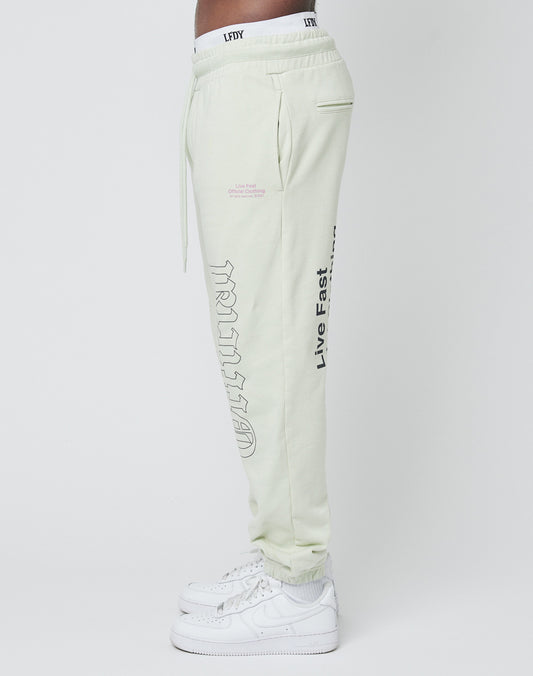 A person wears light-colored LFDY Official Jogger Regular Fit pants made from 100% Baumwolle with Live Fast and stylized lettering printed on front and back, paired with white sneakers, shown in side profile against a plain background.