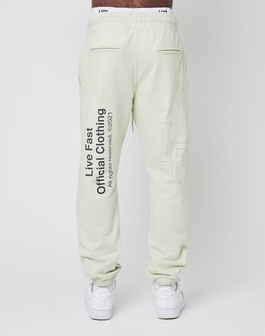 A person wearing light green LFDY Official Jogger Regular Fit pants made from 100% cotton, with "Live Fast. Official Clothing. All rights reserved 2021" on one leg, and white sneakers, shown from the back against a plain background.