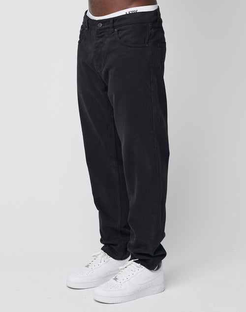 A person wearing LIVE FAST DIE YOUNG Regular Denim, featuring a loose fit and tapered leg, paired with white sneakers and standing against a plain white background.