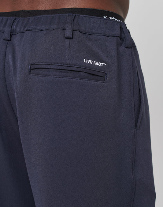 Close-up of the back and side of Regular Casual Pants by LIVE FAST DIE YOUNG in navy blue, featuring a welt back pocket with “LIVE FAST™” embroidered above it in white and an elastic waistband, worn by a person with medium brown skin.
