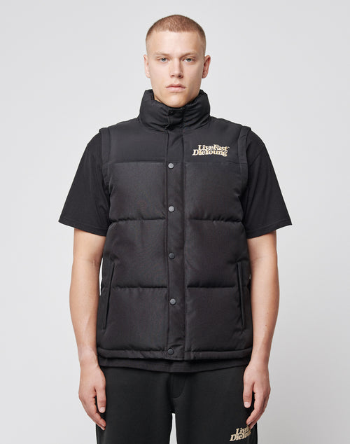 A person with short blond hair wears the LIVE FAST DIE YOUNG Basic Vest (Regular Fit, black, “Life’s Fast. Die Young.” logo) over a black T-shirt and pants, standing facing forward against a plain light background.