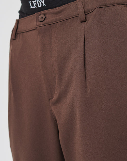 Close-up of a person wearing LIVE FAST DIE YOUNG Regular Casual Pants in brown, featuring a pleated front, button closure, and a black elastic waistband with white LFDY lettering.