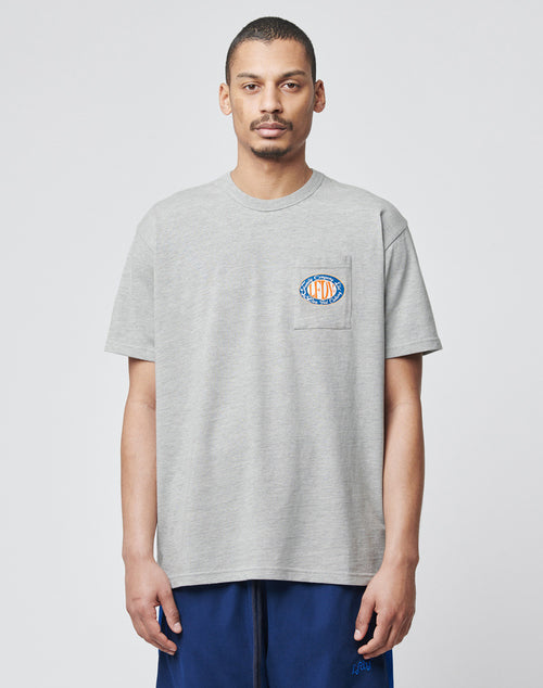 A man with short hair and a trimmed beard wears the LIVE FAST DIE YOUNG Family Pocket Tee, a light gray regular fit T-shirt aus 100% Baumwolle with a small colorful logo on the Brusttasche, paired with dark blue pants. He faces forward against a light background.