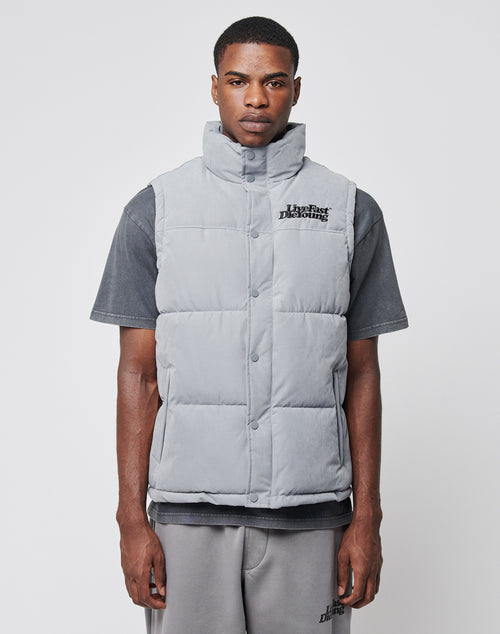 A man wears a LIVE FAST DIE YOUNG Basic Vest in light gray polyester (regular fit) over a dark gray t-shirt and gray shorts, standing against a plain white background and facing forward with a neutral expression.