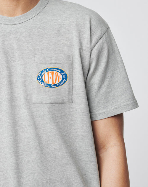 A person wears the LIVE FAST DIE YOUNG Family Pocket Tee, a light gray, regular fit t-shirt made from 100% Baumwolle with a small colorful oval “LFDY” logo and cursive text on the chest pocket. Only upper torso and left arm are visible.