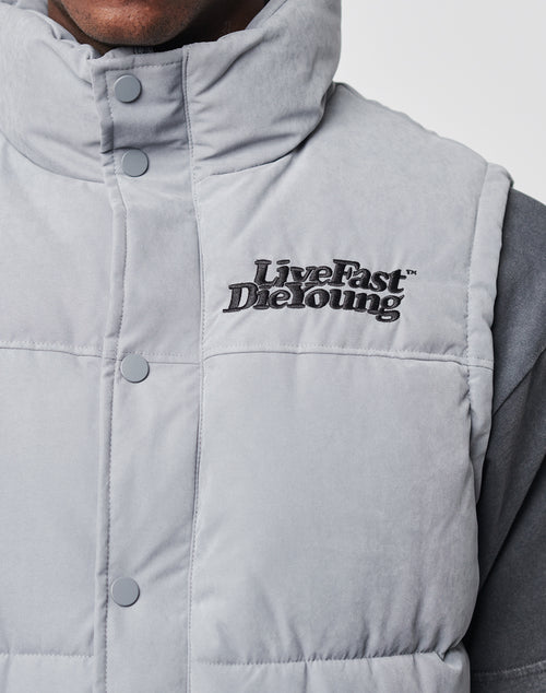 Close-up of a person wearing the Basic Vest by LIVE FAST DIE YOUNG, a light gray regular fit puffer vest with snap buttons and black Live Fast Die Young embroidery on the chest. Part of the persons face and shoulder are visible. Made from durable polyester.