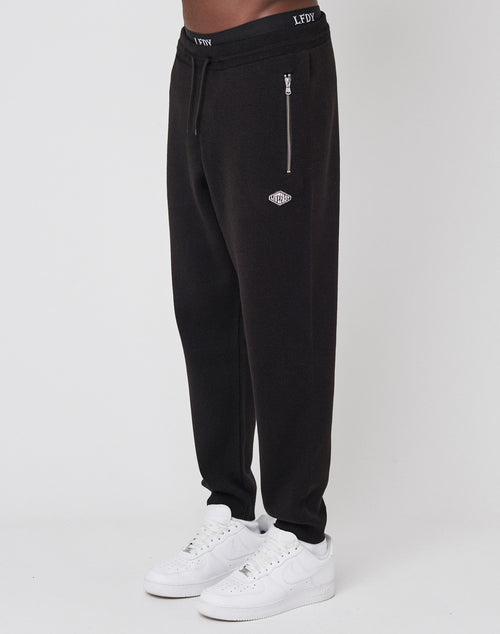 A person wears LIVE FAST DIE YOUNG Knit Jogger pants with an elastic waistband, zippered pocket, and white logo patch, paired with white sneakers, standing against a plain white background.