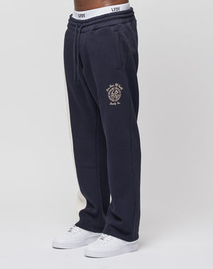 A person wears LIVE FAST DIE YOUNGs LFDY Split Jogger—navy blue with a beige side stripe, embroidered crest and text on the thigh, wide fit, and elastic waist—paired with white sneakers against a plain white background.