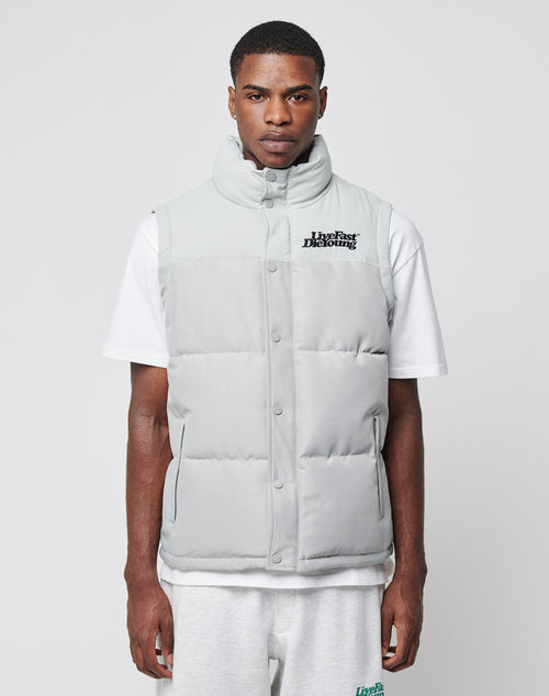 A man stands against a plain background wearing a light grey LIVE FAST DIE YOUNG Basic Vest made from a sleek polyester blend over a white t-shirt and light grey shorts, featuring subtle branding on the left chest.