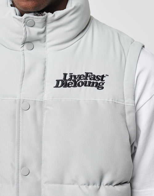 A close-up of a person wearing the LIVE FAST DIE YOUNG Basic Vest in light gray with Live Fast Die Young embroidered in black on the chest and snap buttons, over a white shirt. Only part of their face and upper body are visible.