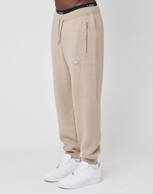 A person wears LIVE FAST DIE YOUNG Knit Jogger sweatpants in beige with a logo patch, regular fit, zippered pocket, and elastic waistband, paired with white sneakers, standing against a plain white background.