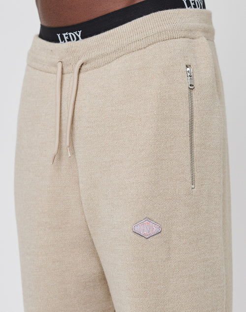 Close-up of a person wearing beige Knit Jogger sweatpants by LIVE FAST DIE YOUNG with an elastic waistband, drawstring, zippered pocket, and small embroidered patch on the thigh. Black “LEDY” underwear peeks above the waist.