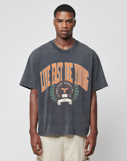A young man stands against a plain background wearing the Live Fast Die Young Tee by LIVE FAST DIE YOUNG, an oversized faded black shirt with bold orange LIVE FAST DIE YOUNG print, laurel wreath, trophy graphic, and ATHLETIC STREET below.