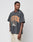 A man models the LIVE FAST DIE YOUNG Live Fast Die Young Tee, a loose, faded black T-shirt with vintage Druck and orange-yellow collegiate text, styled with light beige cargo pants for a relaxed look against a white background.