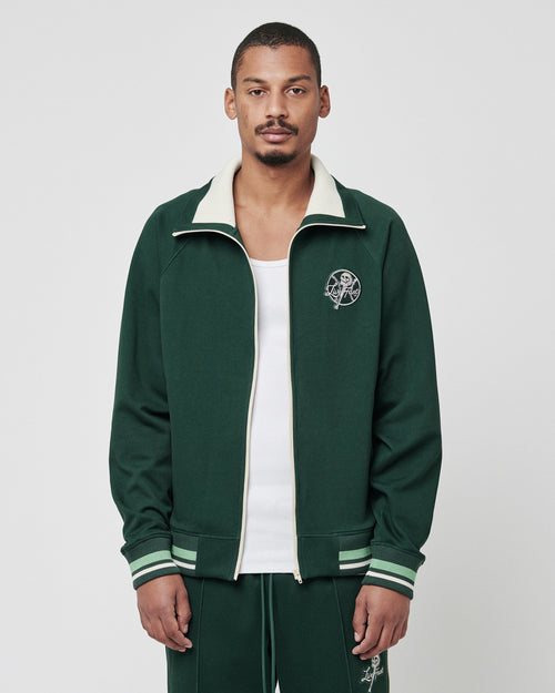 A man stands front-facing in a Regular Fit green Track Suit Jacket with stand-up collar and white trim by LIVE FAST DIE YOUNG, over a white tank top. The logo-embroidered jacket matches his green track pants. The background is plain light grey.