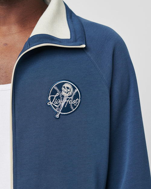 A person wears a blue LIVE FAST DIE YOUNG Track Suit Jacket (Regular Fit) with a white inner collar, white shirt underneath, and features an embroidered skull over crossed baseball bats with Live Fast. Made from polyester baumwolle.