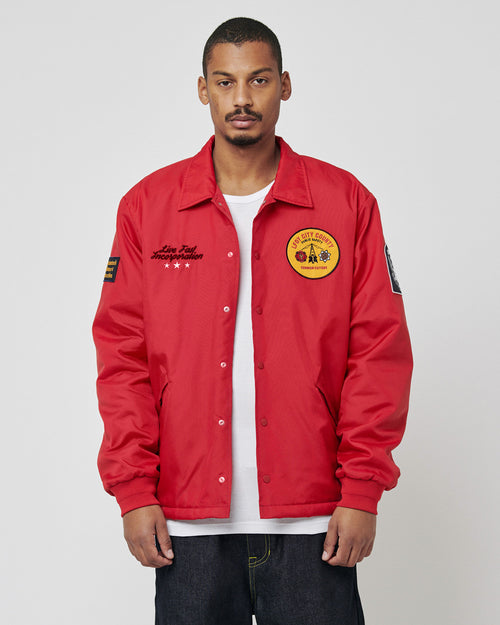 A person wears a bright red LIVE FAST DIE YOUNG LFDY Coach Jacket, regular fit and lightly padded, with patches and embroidery—including a large yellow patch on the left chest—standing forward against a plain light gray background.