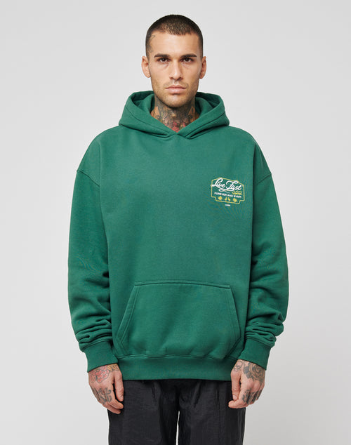 A man with short hair and tattoos wears the LIVE FAST DIE YOUNG Live Fast Greetings Hooded sweatshirt in green, oversized fit, with a small yellow chest graphic. Made from Baumwolle Polyester, he stands relaxed against a plain background.