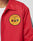 A person wearing a red LFDY Coach Jacket by LIVE FAST DIE YOUNG with a yellow patch reading LEVY CITY COUNTY PUBLIC SAFETY COMMUNICATIONS, featuring fire and police symbols.