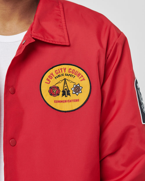 A person wearing a red LFDY Coach Jacket by LIVE FAST DIE YOUNG with a yellow patch reading LEVY CITY COUNTY PUBLIC SAFETY COMMUNICATIONS, featuring fire and police symbols.