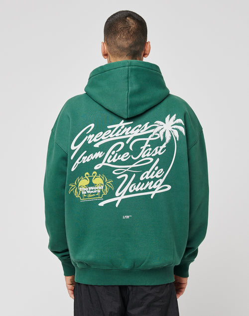 Someone is seen from behind wearing the LIVE FAST DIE YOUNG Live Fast Greetings Hooded, an oversized green hoodie featuring white script text—Greetings from Live Fast die Young—palm tree graphics, and yellow accent text on the back.