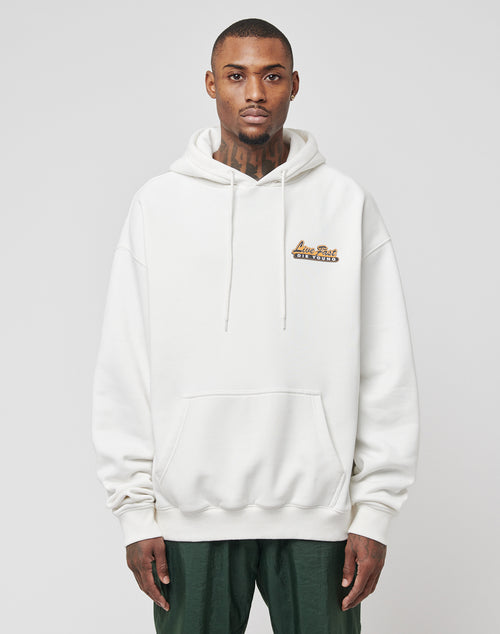 A man with short hair and neck tattoos stands facing forward against a light gray background, wearing dark green pants and a white LIVE FAST DIE YOUNG Home Protection Hooded sweatshirt with a small logo on the left chest.