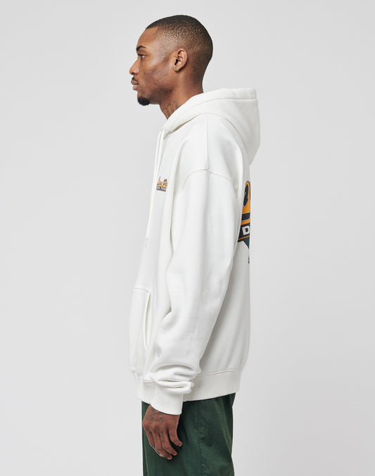 A man stands in profile wearing the LIVE FAST DIE YOUNG Home Protection Hooded, an oversized white hoodie with drawstring hood, graphics and text on the back and left chest, paired with green pants against a plain light gray background.