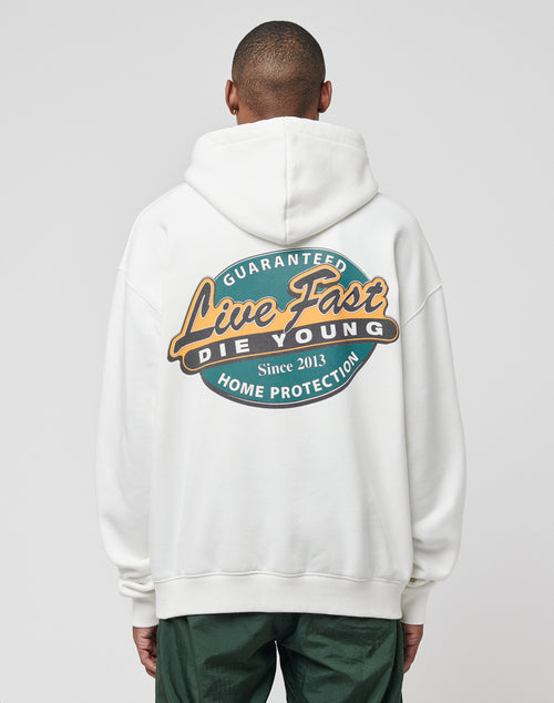 A person wearing the Home Protection Hooded by LIVE FAST DIE YOUNG—an oversized white Baumwolle Polyester hoodie with a large back graphic: Guaranteed Live Fast Die Young Since 2013 Home Protection. Only their back is visible.