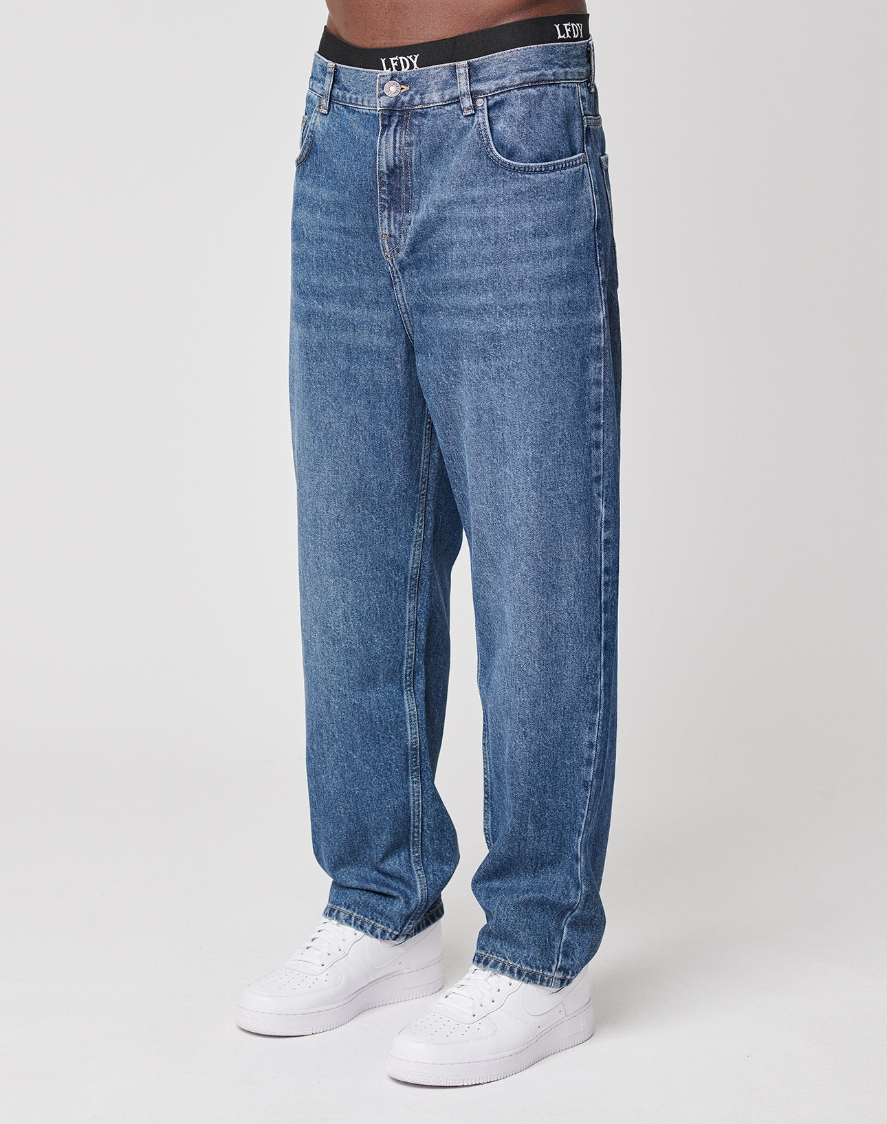 A person wears loose-fitting LFDY Wide Denim jeans in blue with a black waistband and white sneakers, standing against a plain white background.