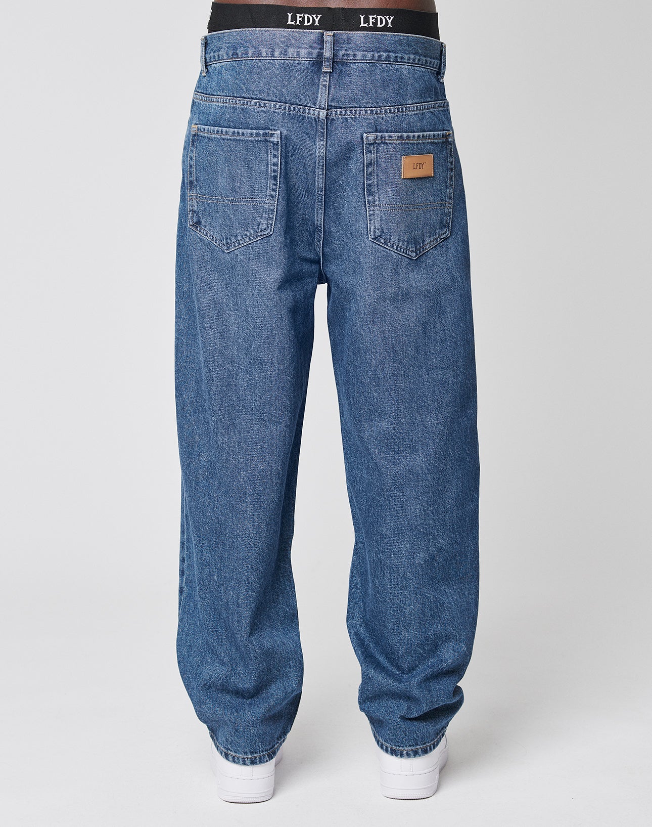 Someone wearing loose-fitting, blue LFDY Wide Denim jeans with a black LFDY waistband. Made from 100% cotton, the jeans feature two large back pockets (one with a brown leather patch), paired with white sneakers.