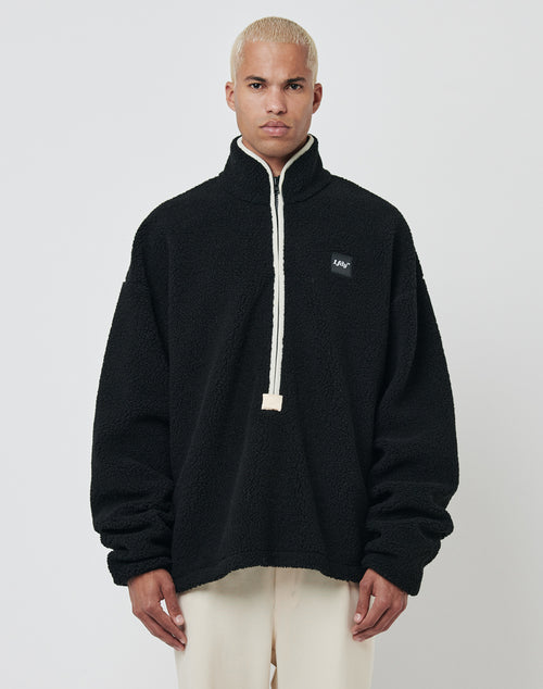 A person with short bleached blonde hair wears the LFDY Teddy Half Zip fleece in black with a high collar and off-white drawstrings, paired with cream pants, standing against a plain light gray background.