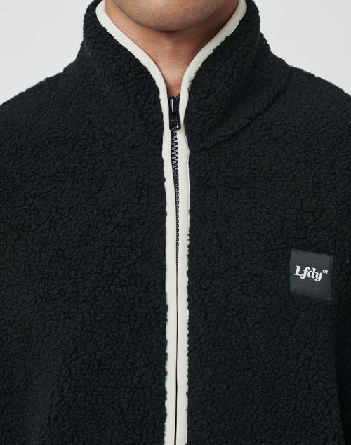 A close-up of a person wearing the LFDY Teddy Half Zip, a black sherpa fleece jacket with a white zipper lining and LFDY logo patch on the chest. The half zip and oversized fit create a relaxed, modern look. The person’s face is partially out of frame.