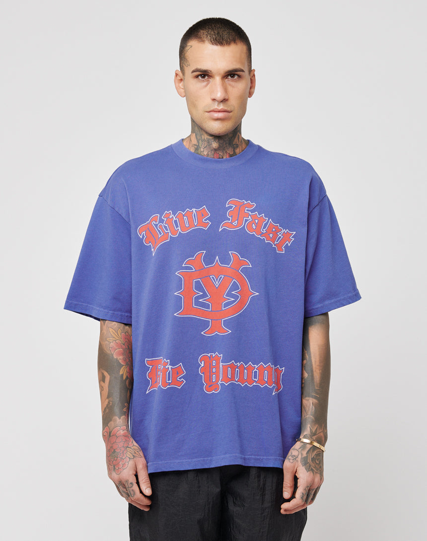 A tattooed man wears the LIVE FAST DIE YOUNG DY Tee, a regular fit made from 100% cotton, with bold red and white Live Fast Die Young text and graphic. Its vintage effect stands out as he faces forward against a plain white background.