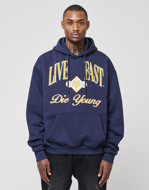 A man wears the LIVE FAST DIE YOUNG Live Fast Campus Hooded, an oversized navy hoodie with a doppellagige Kapuze and yellow and white “LIVE FAST Die Young” text, standing against a plain white background.