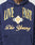 A person wearing the Live Fast Campus Hooded by LIVE FAST DIE YOUNG: a navy blue oversized hoodie with bold yellow LIVE FAST, cursive Die Young, baseballs, a diamond design, and a trendy vintage effect.