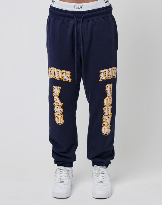 Someone wearing the Live Fast Sweat Jogger by LIVE FAST DIE YOUNG, a regular fit navy blue pant made from 100% cotton, featuring bold “LIVE FAST” and “DIE YOUNG” text in yellow and white, paired with white sneakers.