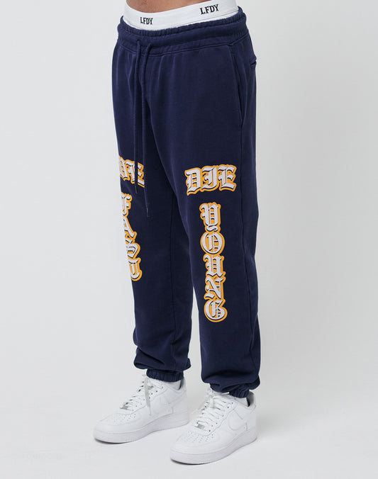Someone wears the Live Fast Sweat Jogger by LIVE FAST DIE YOUNG in navy blue (Regular Fit), featuring bold yellow DIE YOUNG text on the legs, an LFDY-branded waistband, 100% Baumwolle, paired with white sneakers. | model