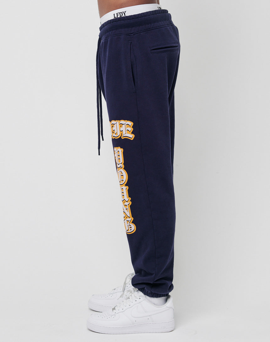 A person wears LIVE FAST DIE YOUNG's Live Fast Sweat Jogger in Regular Fit navy blue 100% Baumwolle, with yellow Old English lettering down the left leg, white sneakers, and a visible white waistband on a plain white background.
