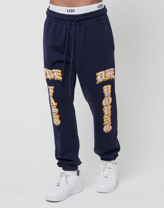 A person wearing navy blue Live Fast Sweat Jogger pants by LIVE FAST DIE YOUNG, made from 100% Baumwolle, with "DIE FAST" and "DIE YOUNG" in yellow on the legs, paired with white sneakers.