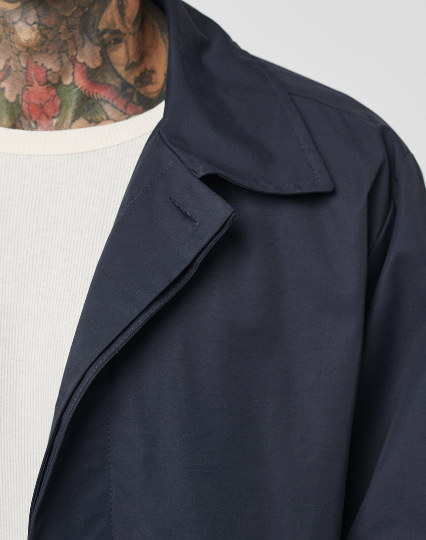 Close-up of a person in a navy LFDY Trench Coat by LIVE FAST DIE YOUNG, layered over a white ribbed shirt. Colorful floral tattoos are visible on the neck and part of the face. Background is plain and light-colored.