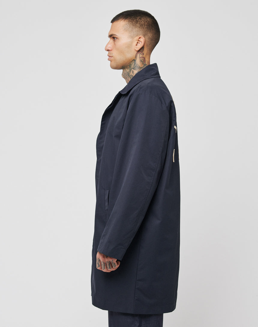 A man with tattoos on his neck and hand stands in profile, wearing the dark navy LFDY Trench Coat by LIVE FAST DIE YOUNG, featuring a concealed button placket, set against a plain light gray background.