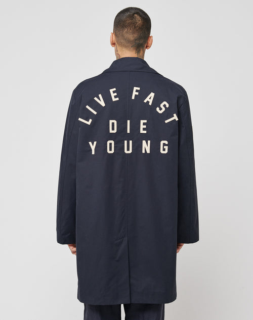 A person with short hair is seen from behind in a regular fit LFDY Trench Coat by LIVE FAST DIE YOUNG, featuring bold white "LIVE FAST DIE YOUNG" lettering on the back, set against a plain light background.