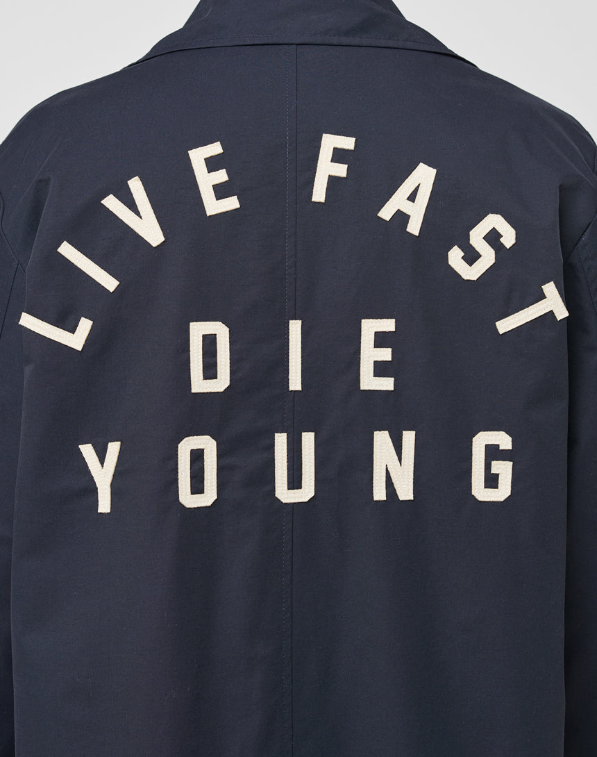 The LFDY Trench Coat by LIVE FAST DIE YOUNG is a dark regular fit coat with bold white "LIVE FAST DIE YOUNG" lettering in three lines on the back and a verdeckte Knopfleiste for a sleek look.