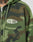A close-up of the LFDY Army Hooded Zip by LIVE FAST DIE YOUNG in green camouflage, featuring LIVE FAST in white on the left chest. A tattooed neck and chin appear above the hoodie, emphasizing its cool vintage effect.