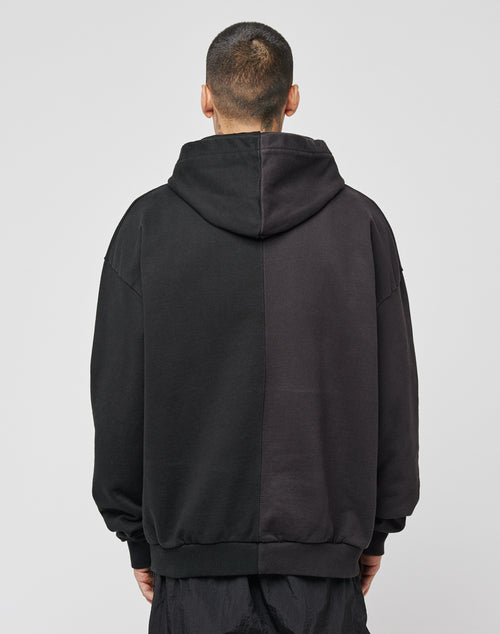 A person with short hair faces away, wearing the LFDY Split Hooded—an oversized cotton hoodie divided vertically into two dark shades, hood up and arms at their sides, set against a plain light background.
