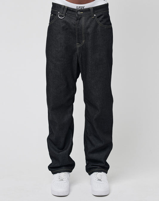 A person wears LIVE FAST DIE YOUNG’s Work Denim—loose-fitting, dark blue raw-denim jeans with visible stitching—paired with white sneakers and a white shirt. The relaxed-fit jeans bunch slightly at the ankles. Background is plain white.