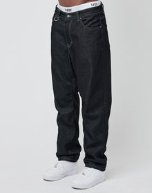 Someone wears loose-fitting Work Denim jeans by LIVE FAST DIE YOUNG, white sneakers, and shows the waistband of white LFDY underwear. The deep indigo denim stands out against a plain, light backdrop.